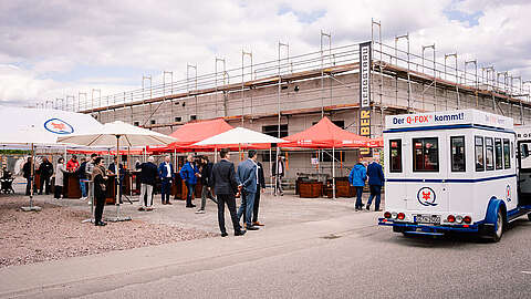 Die Menschen versammelten sich bei einer Veranstaltung im Freien in der Nähe einer Baustelle, auf der mehrere Zelte mit Markenlogos und ein weißer Werbe-Shuttlebus im Vordergrund standen.