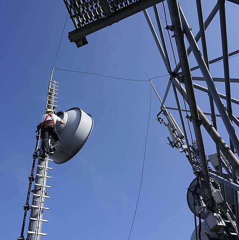 Ein Techniker klettert die Richtfunkantenne hoch. Im Hintergrund sieht man den Himmel