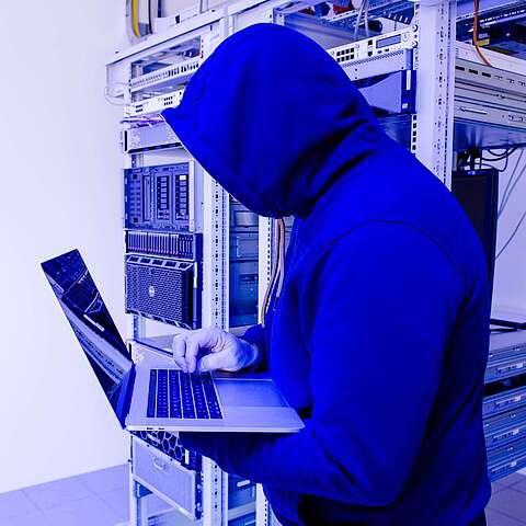 Ein verhüllter Mann im Kaputzenpulli steht in einem Serverraum, einen Laptop benutzend.