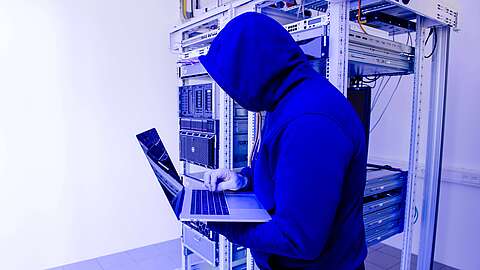 Ein verhüllter Mann im Kaputzenpulli steht in einem Serverraum, einen Laptop benutzend.